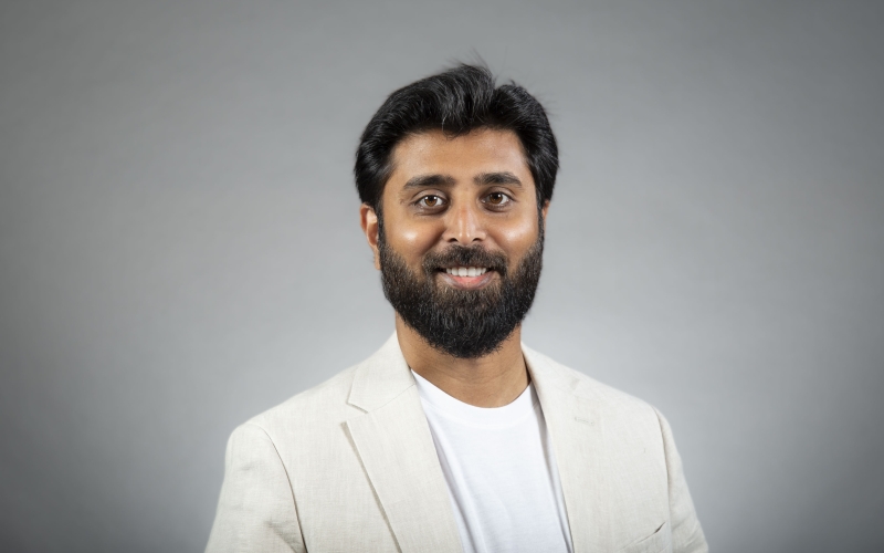 A man with dark brown hair wearing a white T-shirt with white linen blazer smiles for a portrait against a gray backdrop.