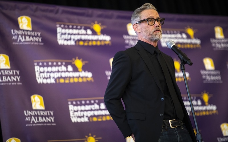 A bearded man with gray/black hair wearing dark-rimmed glasses and wearing all black standing at a microphone in front of a purple backdrop with the words "University at Albany" and "Research and Entrepreneurship Week" on it.