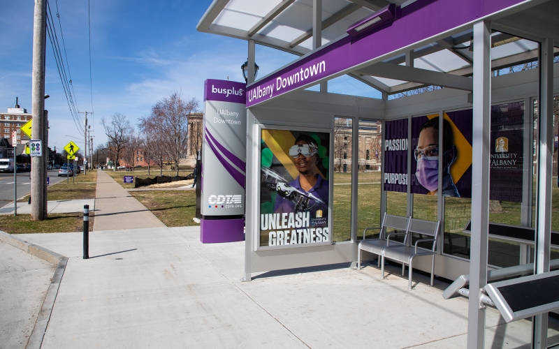 A purple CDTA bus shelter displaying “UAlbany Downtown” and “Unleash Greatness” signage, with metal seating beneath the covered glass structure.