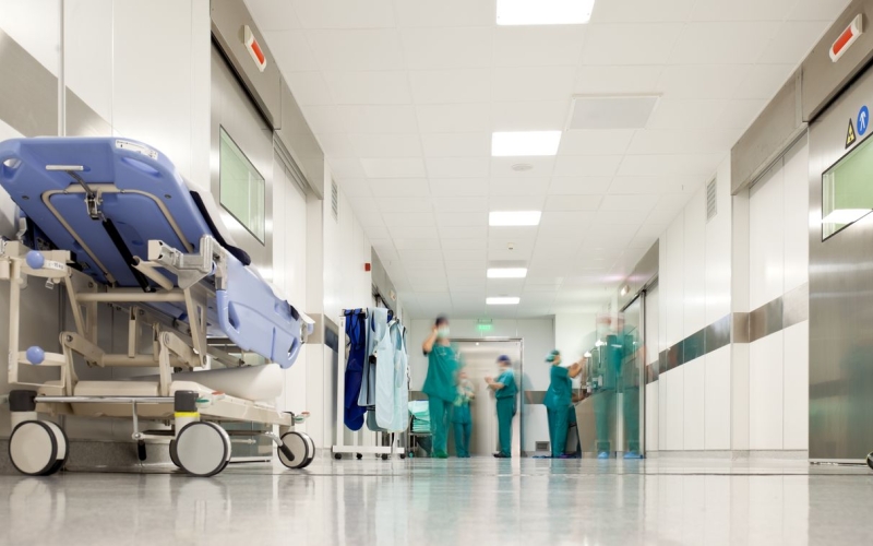 Healthcare workers wearing green scrubs, masks and surgical caps work at the end of a brightly lit hospital hallway.