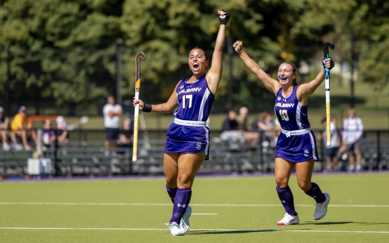 A young woman with dark brown hair in a purple field hockey uniform of the UAlbany Great Danes wearing number 17 lifts her arm in celebration on the turf field.