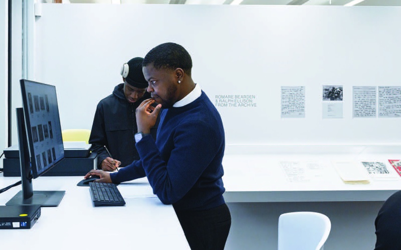 Two students view an archive of materials in a white study space with a sign that reads Romare Bearden & Ralph Ellison: From the Archive on the wall behind them.