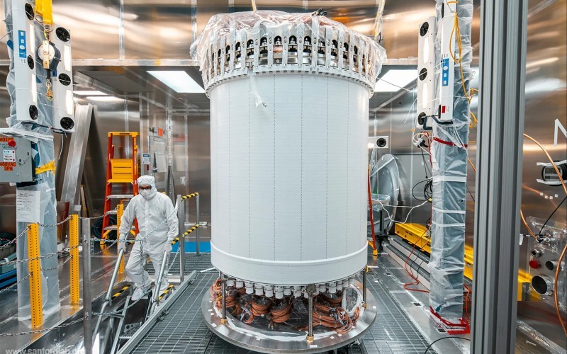 A researcher in protective gear works near a tall, cylindrical scientific instrument with visible wiring. The walls surrounding it are reflective Teflon