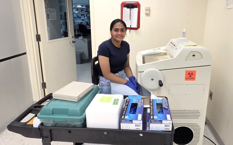 A young woman, seated, wearing blue jeans, a navy T-shirt, and blue latex glove, works at a machine in a biology lab. The machine is made of beige plastic and features a glass window on top and a circular crank on the side. A cart full of lab supplies is in front of her. 