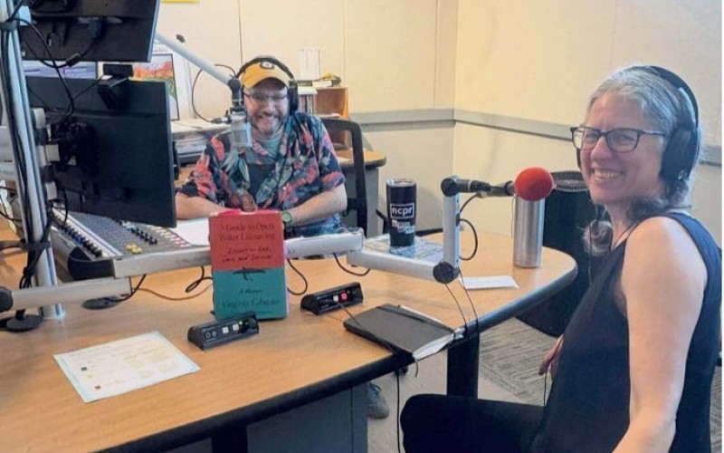 A man and a woman, both smiling, with headphones and microphones turn to face the camera