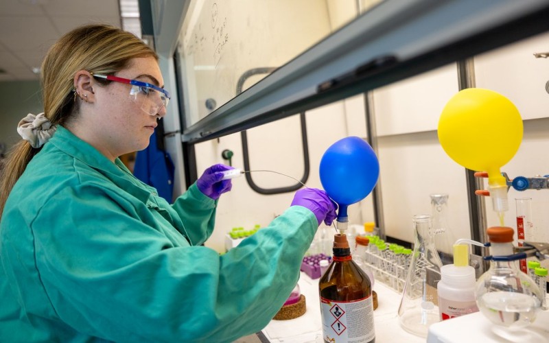 A young woman wearing a green lab coat and purple gloves dips an instrument with a long wire probe into a brown bottle with a small blue balloon attached to its mouth. 