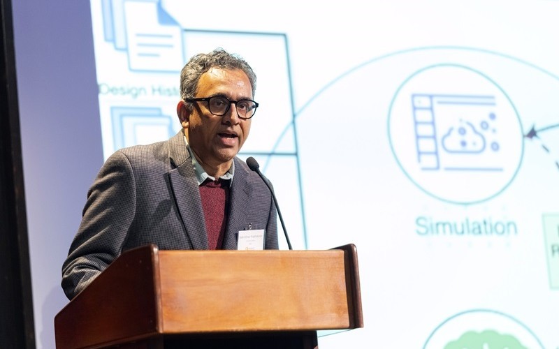 A man with eyeglasses wearing a gray blazer and red sweater stands speaking at a brown lectern with various technical images and the word "Simulation" projected on the screen behind him in blue and green.