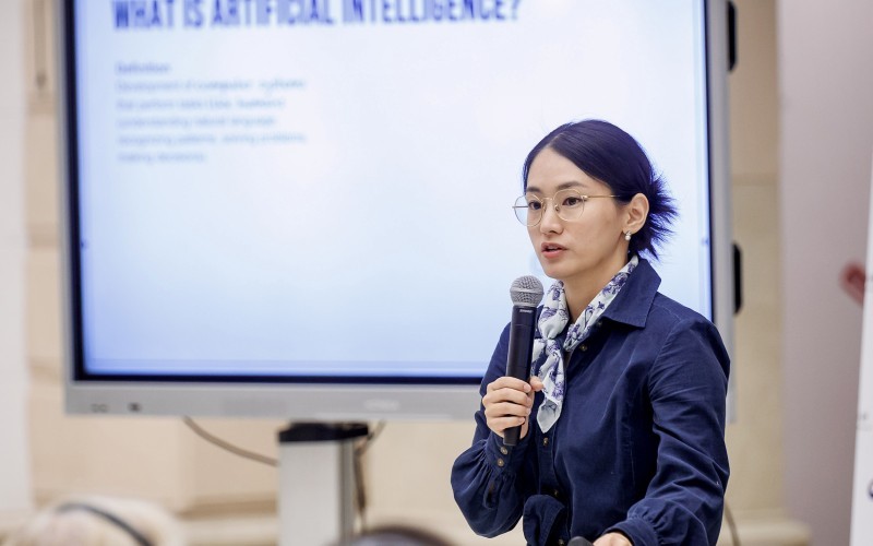 A young woman with dark black hair and glasses wearing a blue blouse and scarf speaks before a monitor with the phrase "What is Aritificial Intelligence?" written on it.