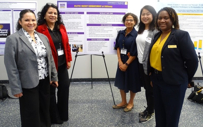 Five women in professional attire stand, smiling, in front of a purple poster set on a black tripod. The poster is titled 'BILPOC Faculty Advancement Initiative at UAlbany' and has subheads reading: who we are, our goals, what we do, outcomes, and our mission."