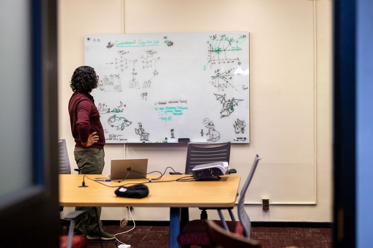 A man wearing a maroon shirt stands facing a whiteboard covered in notes and drawingswith his hands on his hips.