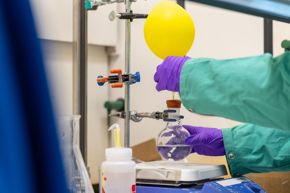 A person wearing purple gloves handles a florence flask with a yellow balloon attached to its mouth.