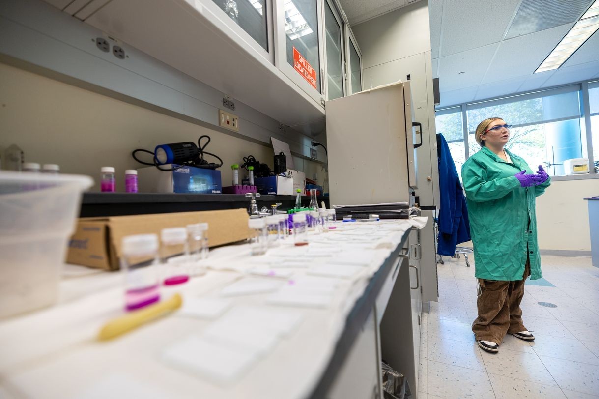 A woman wearing a green lab coat stands beside a lab bench containing an assortment of clear vials containing a purple substance.