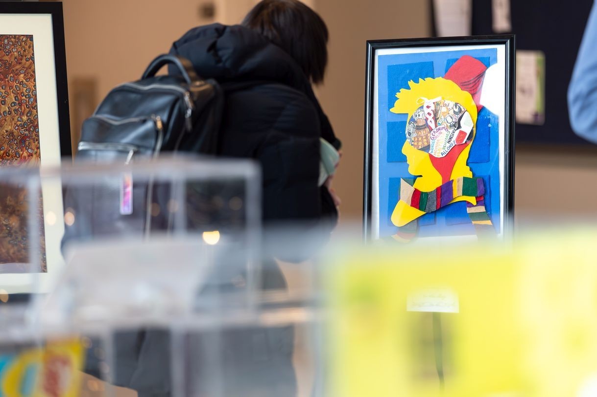 Photo of an artwork set behind display cases arranged in the foreground. The framed artwork features a paper-based collage consisting of a yellow outline of a person‘s head, a colorful band layered as a scarf, and a multicolored brain in the center, set against a blue background. 
