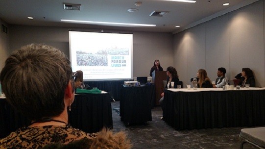 A woman stands at a podium and presents a slideshow to a conference room full of people.