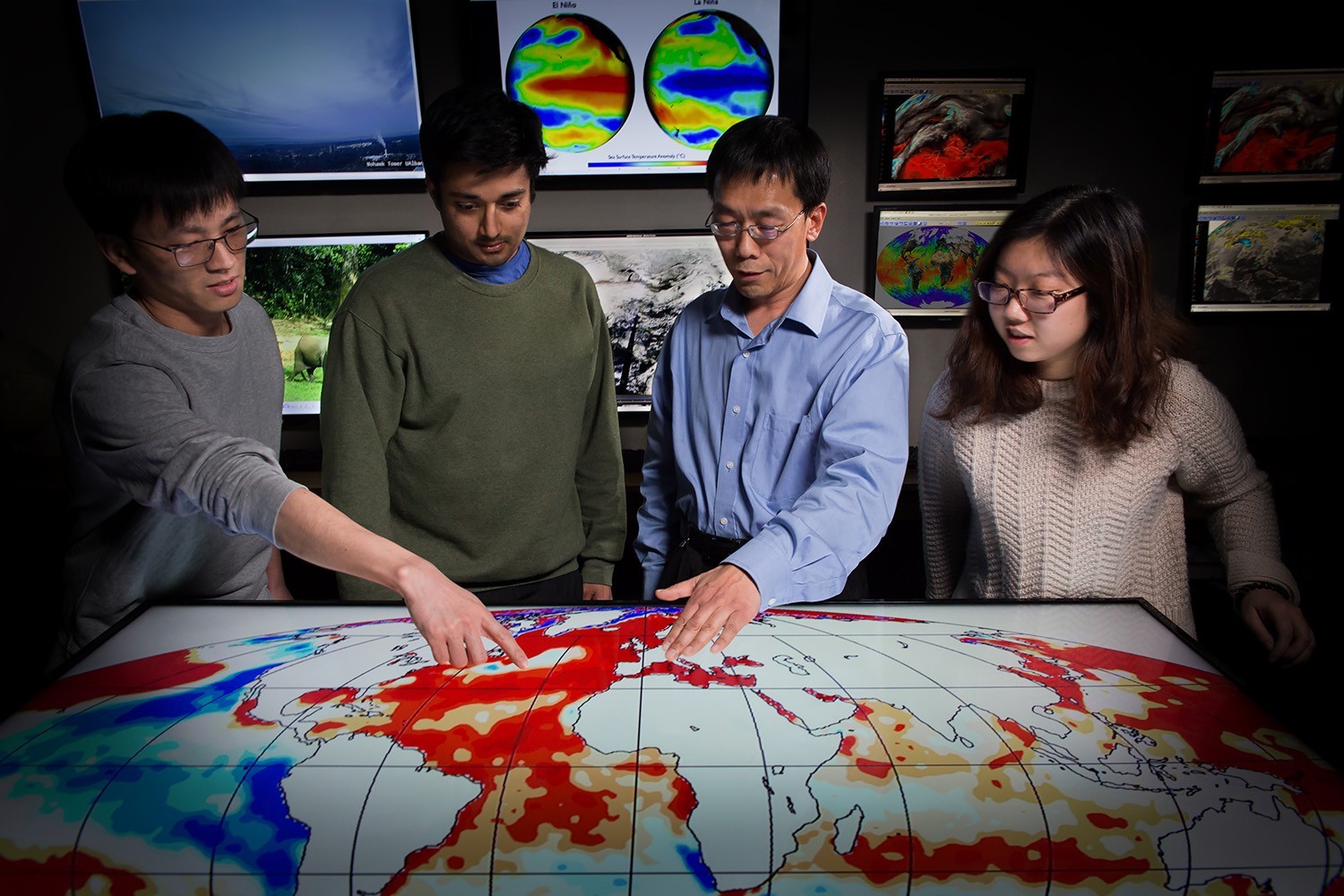4 people looking at a weather map on a table with screens of weather maps behind them