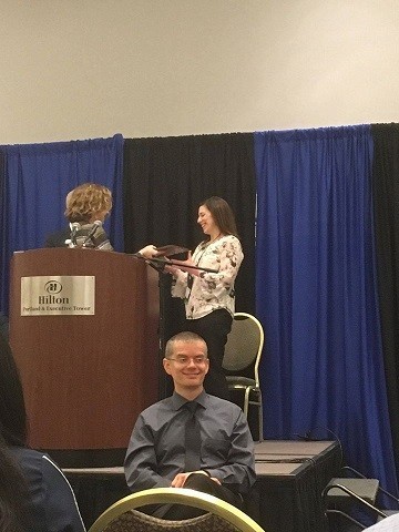 A woman with brown hair and a white patterned blouse accepts a plaque from a woman with short blonde hair behind a podium on stage in front of crowded room.