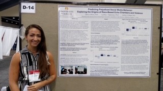 A woman with long brown hair. in a black and white dress wears a name tag and smiles next to a research poster