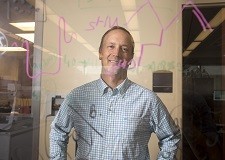 A man in a collared shirt smiles in the background, seen through a glass window with scientific writing on it.