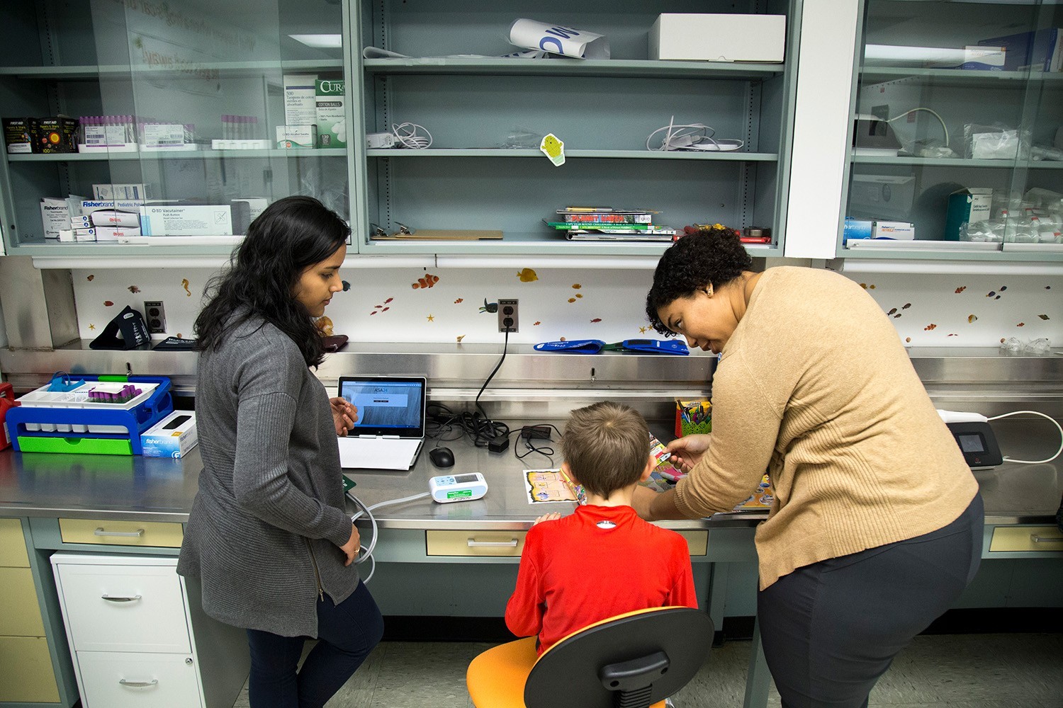 An intern and a professional stand on either side of a child wearing a bright red shirt as he completes a medical survey.