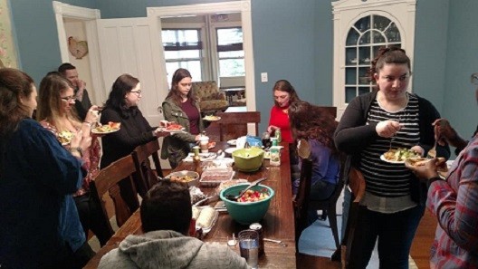 Ten people sit and stand around a dining room table, eating, talking and laughing