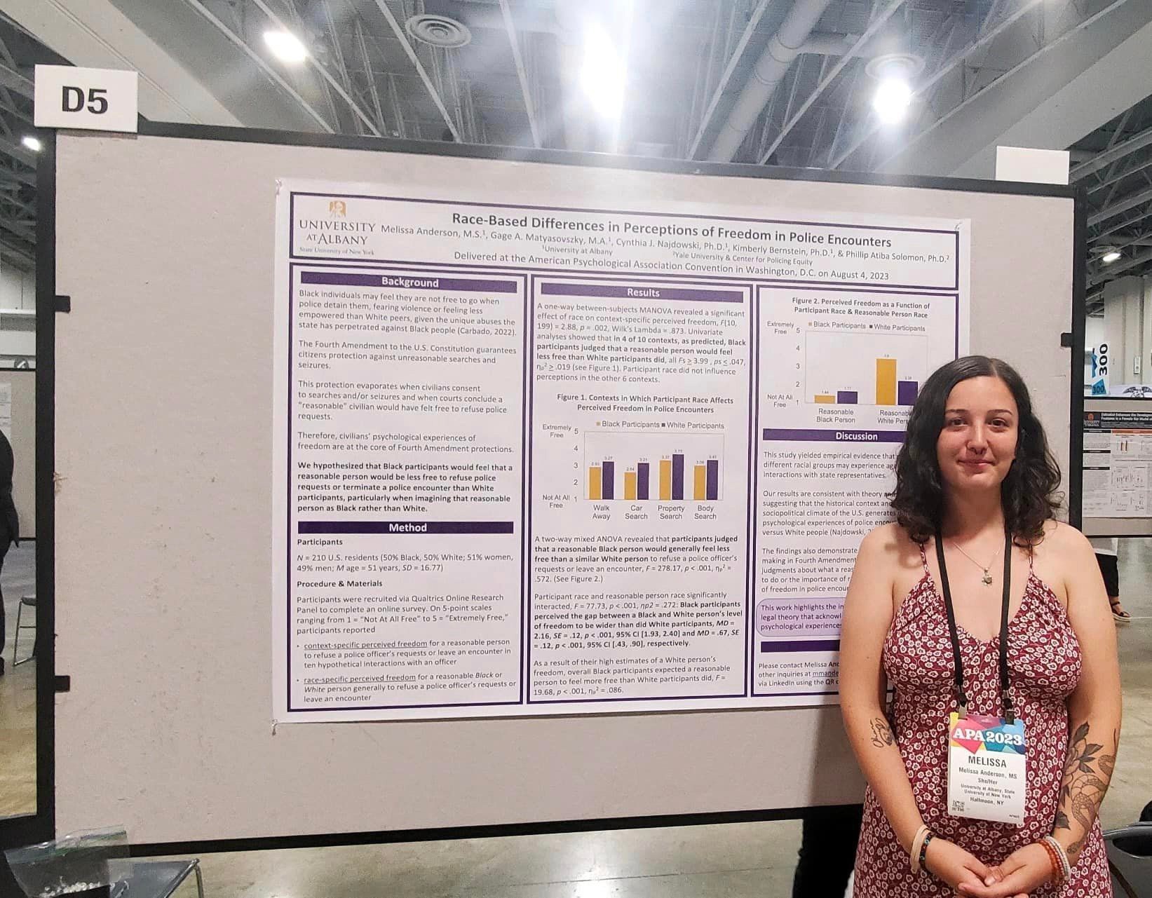 A woman in a red and white floral dress, wearing a lanyard with shoulder-length brown hair stands in front of a research poster titled, 'Raced-Based Difference in Perceptions of Freedom in Police Encounters'