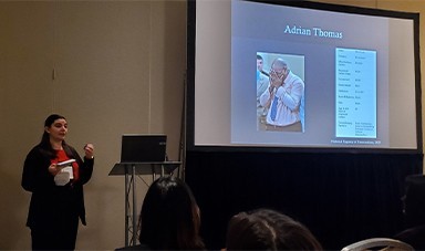 A woman with dark hair in a red shirt and black blazer stands by a podium in front of a room of people while presenting a slideshow. On a screen behind her, a slide reads 'Adrian Thomas' with a picture of a man holding his face in his hands.
