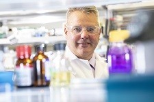 A man in lab coat is photographed through an opening in a lab shelf, framed by bottles and other lab supplies.