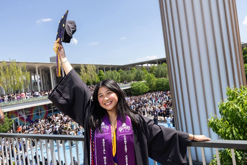 Student holding her mortarboard up in celebration in front of the fountain.
