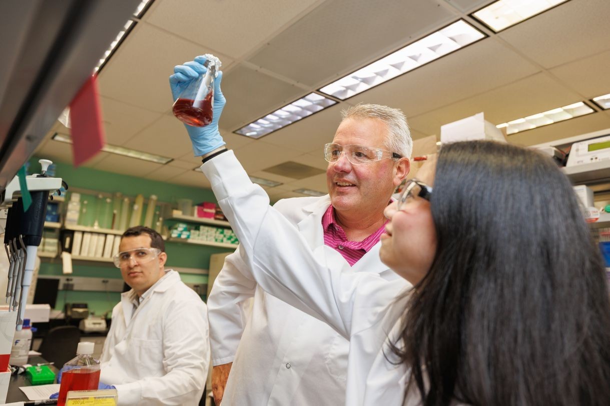 A young woman wearing lab goggles and a white lab coat holds up a beaker containing a red liquid. A man wearing a white lab coat and plastic protective goggles stands behind her, observing the liquid inside the beaker. A man wearing a white lab coat and goggles works at the lab bench behind them. 