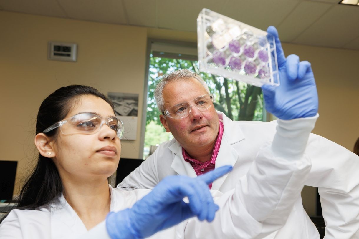 A young woman wearing lab goggles, a white lab coat and purple gloves points to a clear plastic tray containing a dozen smaller circular petri dishes containing small swatches of purple growth media. A man wearing a white lab coat, salmon shirt and clear lab goggles observes the tray she is holding.  