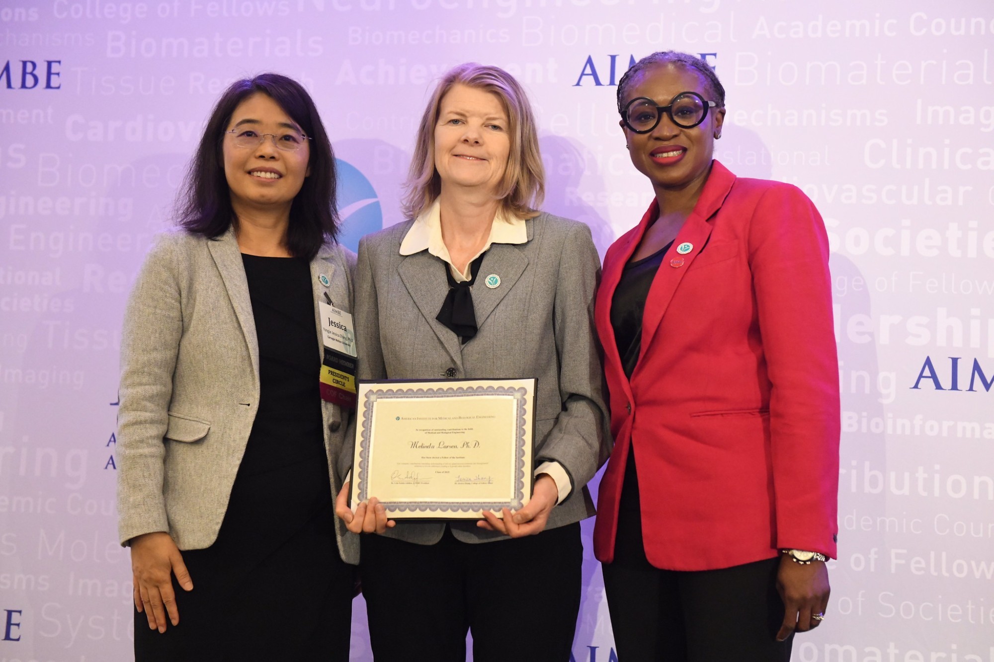  Three women smile for a photo. They are standing against a purple-lit backdrop printed with the name “AIMBE” (in purple) as well as a collection of science-related words in very faint print. The woman at left is wearing rimless glasses, a tan blazer and a black dress. The woman in the center is wearing a gray blazer and black slacks and is holding a certificate. The woman at right is wearing a red blazer and glasses with black circular rims. 