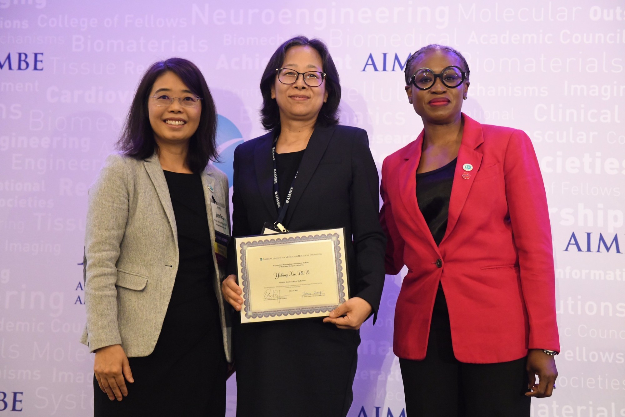 Three women smile for a photo. They are standing against a purple-lit backdrop printed with the name “AIMBE” (in purple) as well as a collection of science-related words in very faint print. The woman at left is wearing rimless glasses, a tan blazer and a black dress. The woman in the center is wearing a black blazer and black square rimmed glasses and is holding a certificate. The woman at right is wearing a red blazer and glasses with black circular rims. 