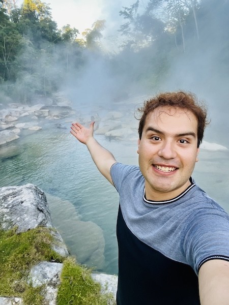 A smiling man stands and gestures toward a steaming river in the jungle