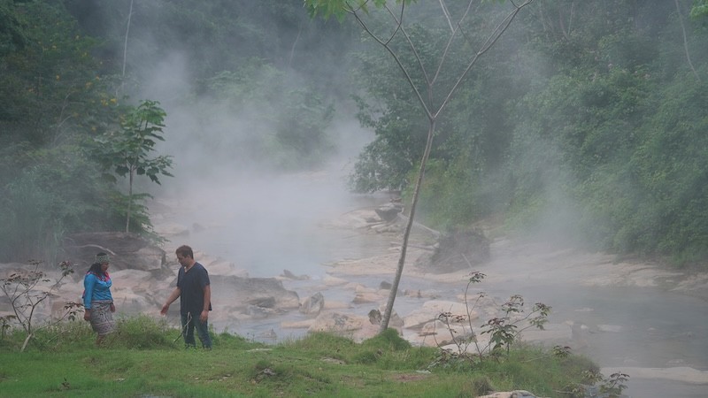 A man and woman stand near a steaming river deep in the jungle