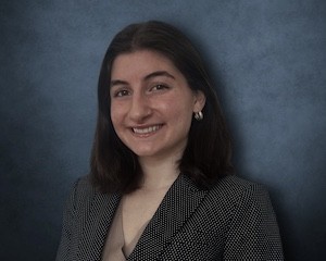 A woman with shoulder-length brown hair wears a blazer and smiles for a portrait against a blue backdrop.