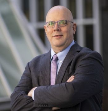 Shawn Bushway in a gray suit and glasses, stands outdoors, arms crossed