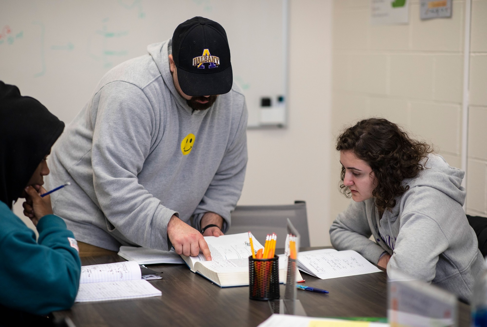 A tutor in a sweatshirt and baseball hat points to a line in a textbook as he leans over a table and two seated students look on.