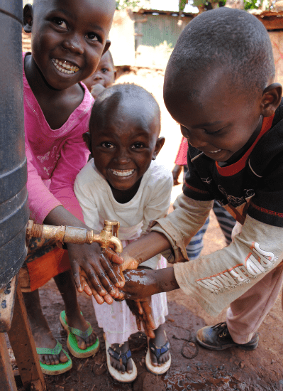 Three children smile while washing their hands outdoors.