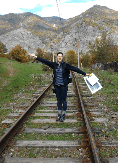 A woman stands on a railroad track in the Country of Georgia, with mountains and grass behind her.