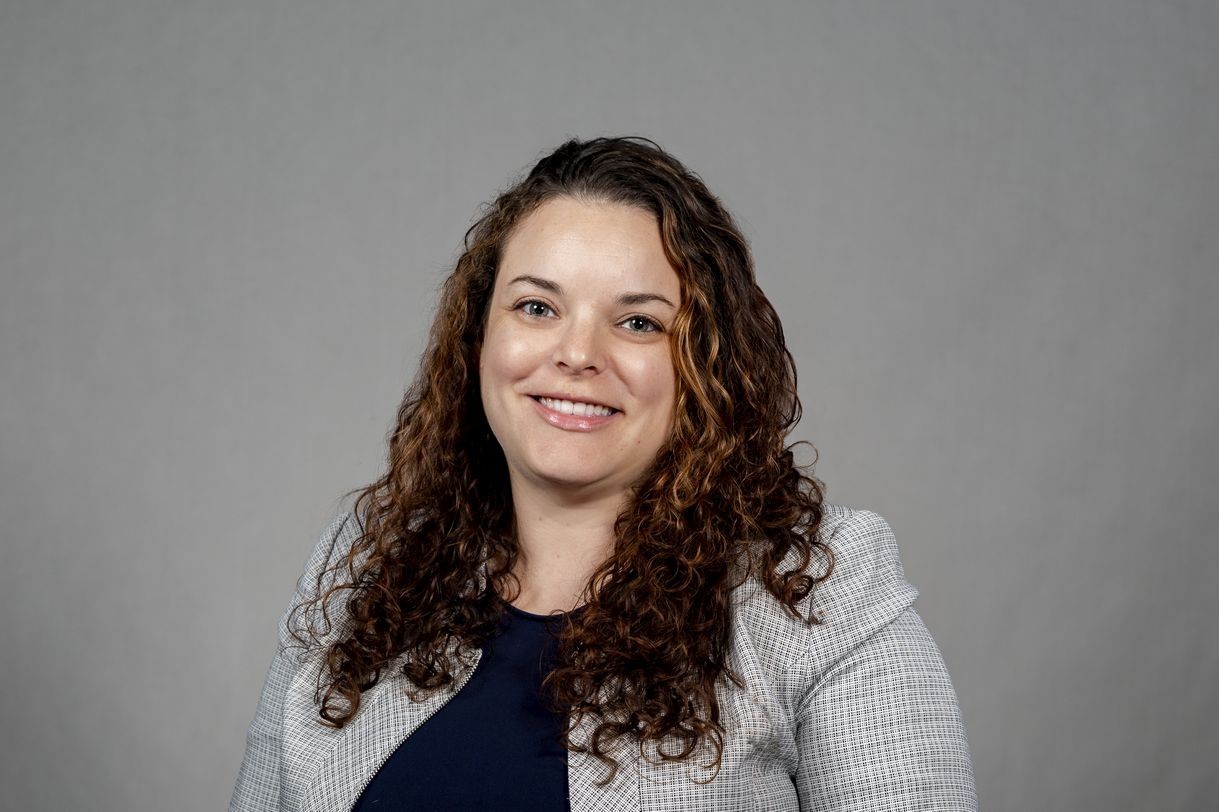 A woman with curly brown hair wearing a gray blazer and navy shirt smiles for a portrait in front of a gray backdrop.