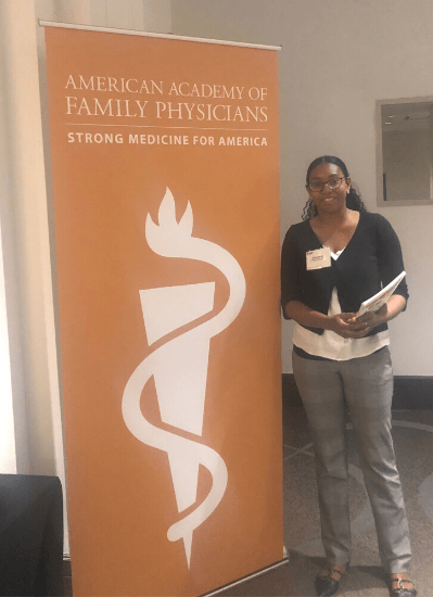 Elizabeth S. stands next to a orange pop-up banner that displays the name of the conference she attended. The conference logo, a white triangle with a squiggle on top, is also on the poster.