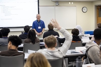 A man looks out over a classroom of high school students, one of whom is raising his hand.
