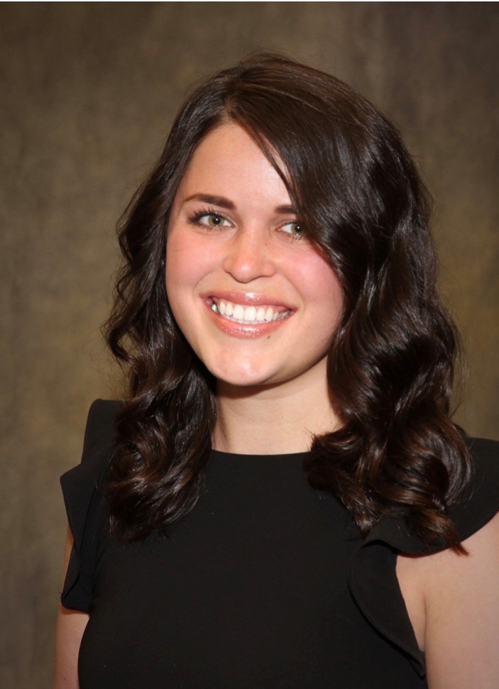 Portrait of Marshall against a brown backdrop. Marshall has long wavy brown hair and is smiling, wearing a black top.