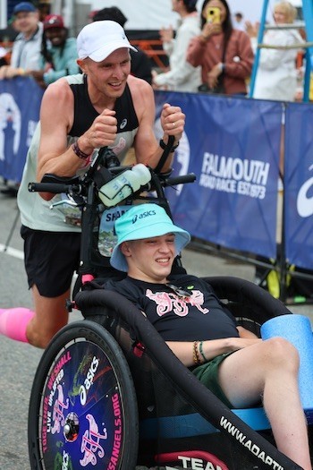 A runner pushes a person in a racing chair along a city street as a crowd of spectators look on.