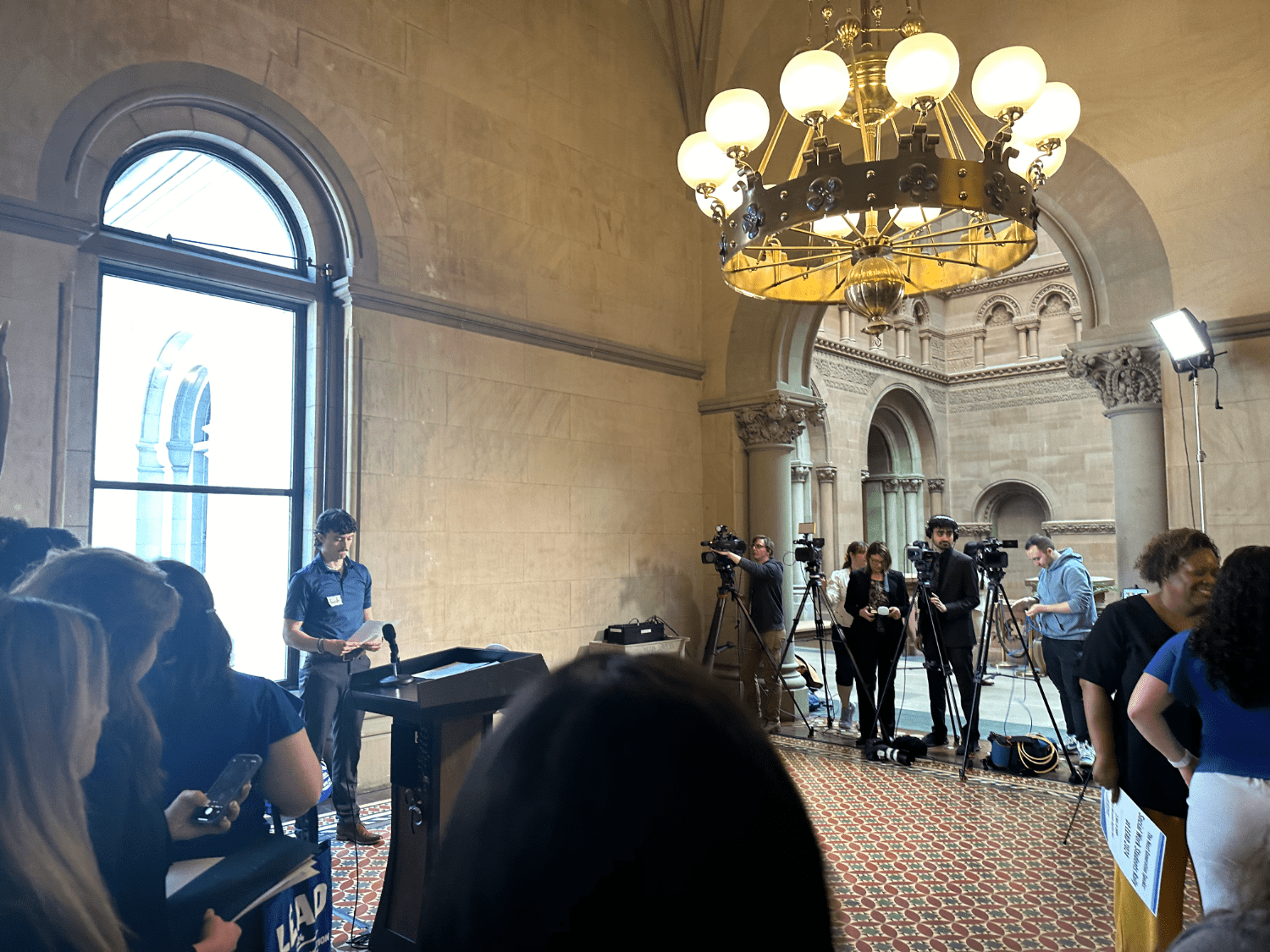 Jacob Schoff speaks at a wooden podium in front of a large arched window in a room in the Capitol. Reporters with camers tripds are in a row at right. A large chandelier hangs overhead and the floor is coated in red and blue tiles. 