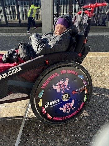 A person bundled up in a thick black coat and purple cap is seated in a racing chair and smiles for a picture.