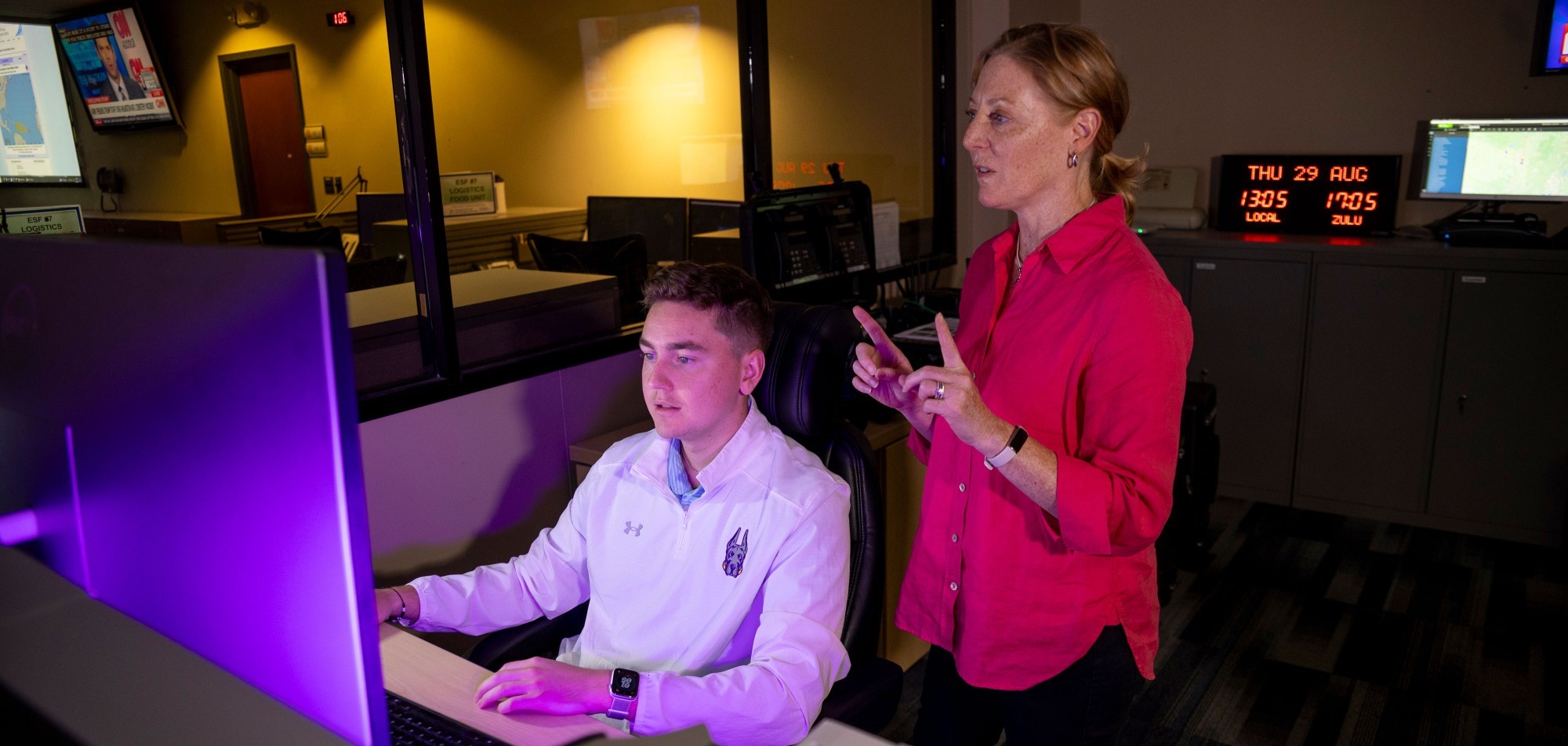 A student intern sits at a desk working on a computer as a supervisor standing beside him provides instruction.