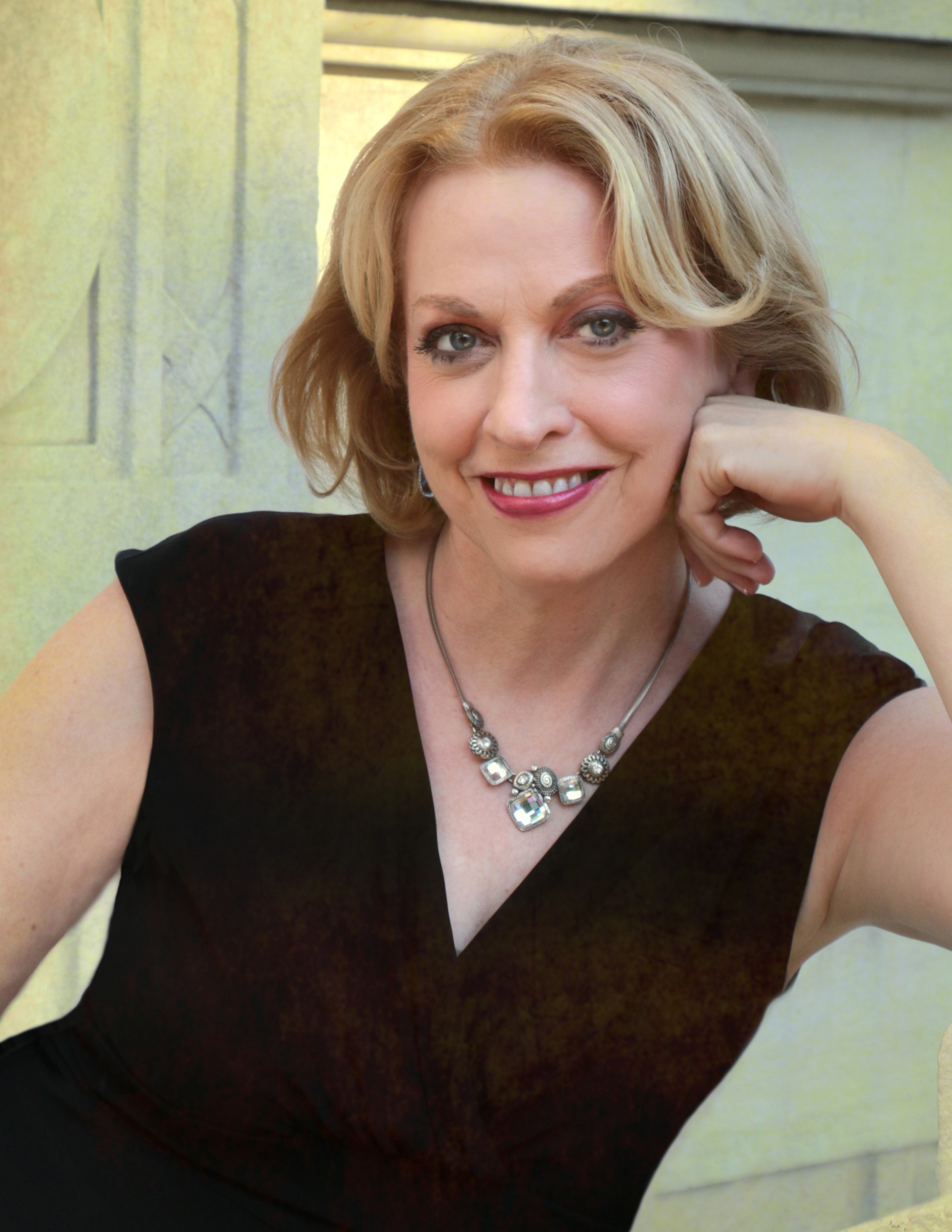 woman with short blonde hair in black dress and silver necklace smiles and leans her cheek on her hand