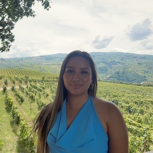 A young woman with long brown hairs wears a blue halter top and smiles for a picture in front of a sweeping green vista.