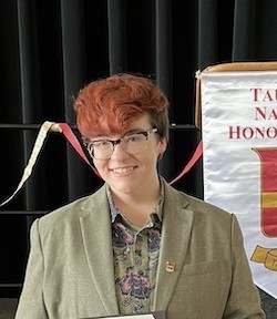 A young person with short red hair and glasses wears a light green blazer and smiles for a portrait.